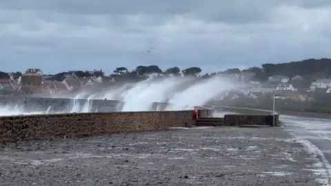 Large waves crash over a seawall in Guernsey during stormy weather brought by Storm Benjamin on Thursday. Parts of the road next to the wall have puddles and surface water. Thick grey clouds are in the sky.
