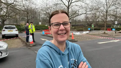 A woman with glasses, brown hair and a blue hoodie crosses her arms and smiles at a camera. She is standing in a car park which has been taped off and turned into a bottled water station. In the background a man with glasses talks to a man wearing a high vis jacket.
