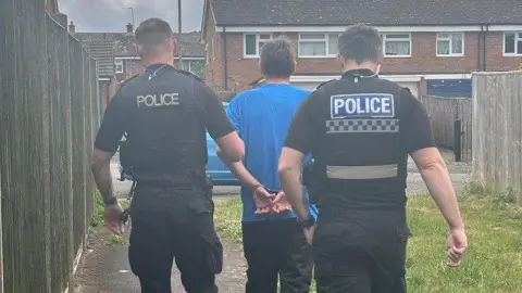 Two Thames Valley Police officers are walking away with a handcuffed man from a yard of a home. They are all with their backs to the camera.