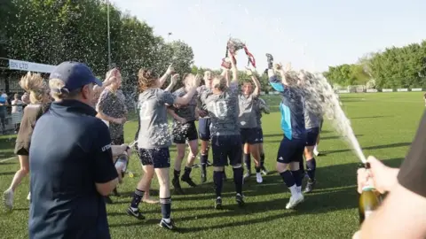 Andrew Harper A group of female footballers celebrate on a grass pitch, spraying champagne
