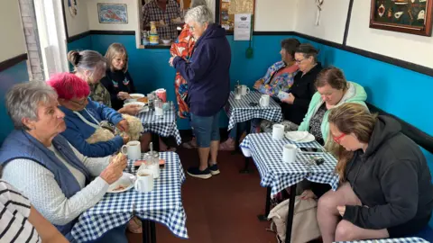 A group of women sitting at a number of tables having a breakfast of bean jar, bacon sandwiches and baps. There is a dog on one of the women's laps.