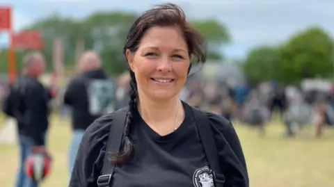 Lisa Brain has dark hair tied in a plait over her right shoulder. She is smiling and wearing a black t-shirt with the black straps of a rucksack over her shoulders. She is standing outside with crowds behind her.