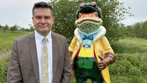 A man in a suit next to a statue of a frog wearing a hat and goggles. The frog statue is also wearing a bow tie and has a camera around his neck. Both are pictured standing in a green field with wild flowers and some trees in the background.