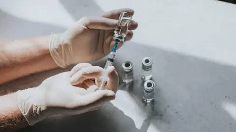 Getty Images A close up shot of gloved hands, filling a needle from a glass vial, there are several other vials on a white table surface