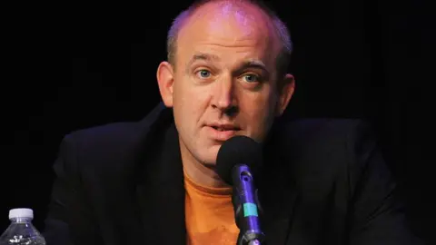 Getty Images Comedian Tim Vine. He is wearning a orange t-shirt with a black jacket over the top. He is speaking into a microphone