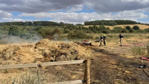 BBC/Suzy Lawson Three firefighters hosing down mounds of straw. There is smoke coming off it. Behind them is a landscape of hills, grass and trees.