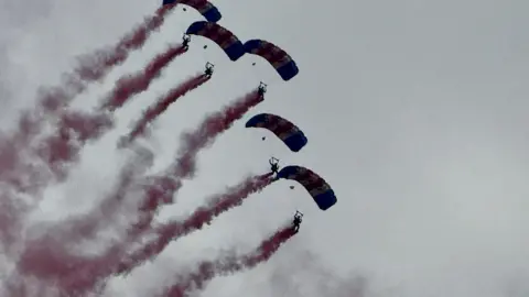 Five people with parachutes deployed falling from a grey sky as red-coloured smoke trails behind each of them.