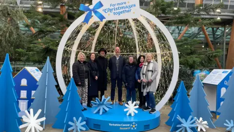 Tom MacDougall/BBC A large, open-front bauble in the Sheffield Winter Gardens, an indoor enclosure filled with trees and plants. It's stood on a blue, Yorkshire Cancer Research branded podium, and surrounded by blue cut-outs of Christmas Trees. Two information boards are positioned either side. Inside the bauble stand a group of representatives from the charity.