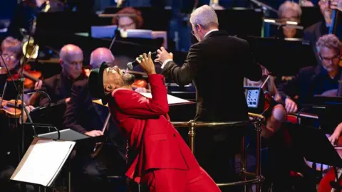 Cad Taylor/BBC Jones leans back singing into his microphone. He wears a bright red coat and black hat. Behind him is a full orchestra