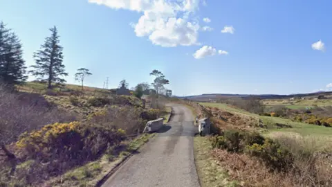 BBC A single-track road and a small bridge with low stone walls. The surrounding landscape is of green fields and areas of gorse.