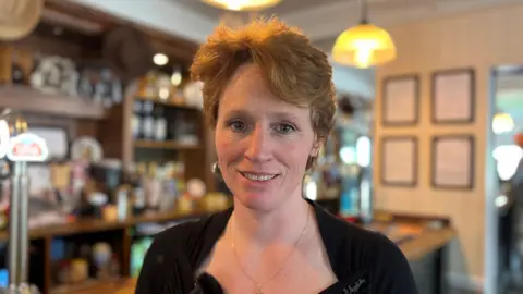 Kerry is wearing a black top and behind a bar in a pub.