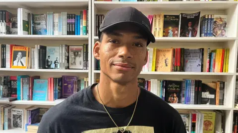 A young man wearing a black baseball cap stands beside some shelves in a bookshop. 