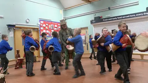 Pop star Tom Morley wears a green top hat with a long green coat. He is standing and banging a drum. He is surrounded by primary school children with drugs. They are in a school hall.