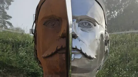 Sculpture showing a face in two halves, one side is shiny silver metal, the other is rust