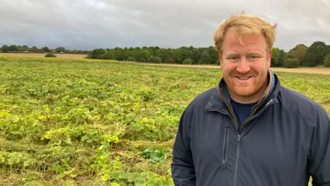 James Grant/ BBC A man in a navy jacket with blonde hair stands in a large squash field.