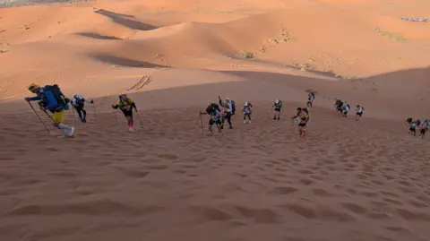 Lenny Worthing People dressed in running clothes hiking up a sand dune in the desert.