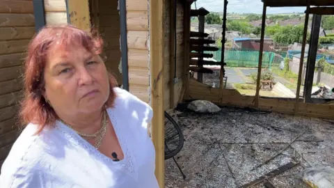 BBC/NATHAN TURVEY A picture of Doreen Smithson who is wearing a white top standing in front of her burnt-out summer house. You can see broken windows, with glass on the floor and charred wood with the allotments in the background.