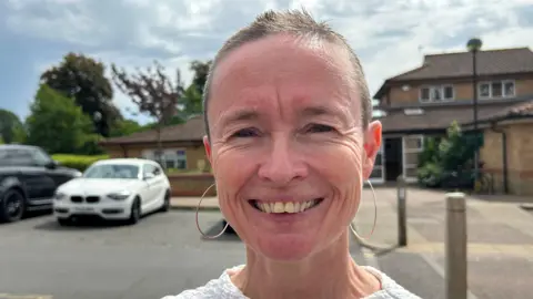 Imogen Lee/BBC Rebecca White is standing in front of a building which has a white door and a path leading up to it. She has short brown hair and is wearing silver hooped earrings and a white T-shirt.