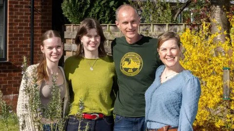 Yorkshire Cancer Research A lady in a blue blouse, from the earlier picture. She stands with a tall bald man with a dark green t shirt. Two girls who appear in their teens or early 20s stand next to them. They all have arms around each other and are smiling at the camera.