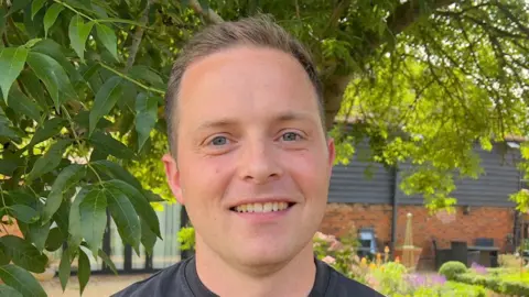 John Fairhall/BBC Tyler Whitnall smiles at the camera. He has short light hair and wears a dark T-shirt.