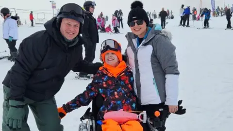 Family photo Brooke Hacker and her parents on a ski slope. Brooke is in a wheelchair wearing a blue, black and orange coat and bright orange hat, scarf and trousers. She has black goggles on her head. Her mother, Rebecca is on her right in a grey and white jacket and a black hat on her head and sunglasses. Brooke's father, Andrew is on her left, in a black coat, black helmet and green trousers.