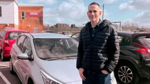 Sion Dafydd is standing outside his car smiling at the camera