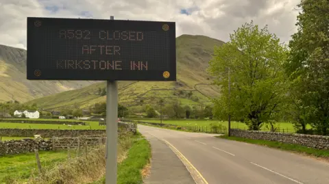 A road warning sign reads A592 CLOSED AFTER KIRKSTONE INN. The sign stands by a beautiful picturesque road in the Lake District with green fells in the background. It is a bright, dry day.