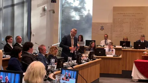 An administrative meeting at Thurrock Borough Council, featuring a man standing and speaking at a central podium surrounded by seated council members and attendees in a wood-panelled chamber.