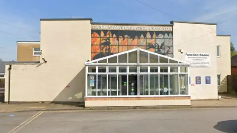 The front entrance to the Lea Hall Miners Welfare Centre. There is a glass windowed front section leading to a stone brick building, painted in ivory, with a mural showing a minor above the entrance.