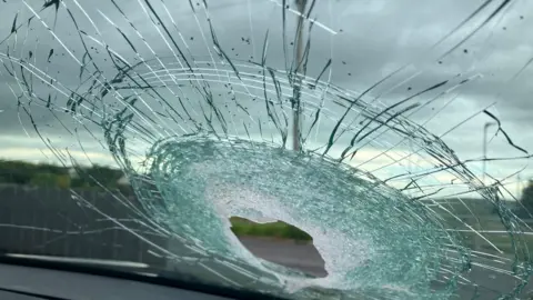 A photo of a smashed up windscreen taken from inside of a car. There is a hole with a lot of damage. The sky is cloudy. A grey fence is in the background. 