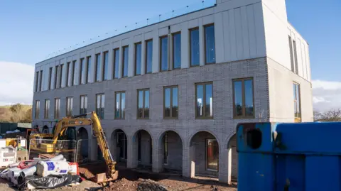 Three storey building with several long windows with white bricks. The sky is blue. There is a yellow digger outside the building. The ground outside the mosque is brown and there is a partially dug hole. There are barricades and construction materials on the left hand side of the image. 