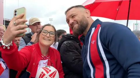 A crowd of rugby fans with player Jared Waerea-Hargreaves. A woman at the front of the crowd is holding a mobile phone up in the air on the left and smiling. She  has oversized black-framed spectacles on her face and is wearing a red bomber jacket over a red and white t-shirt. Her brown hair is tied up in a ponytail. She is holding a red and white rugby ball with the right hand and is standing next to a rugby player, who is smiling at towards the mobile phone. He is wearing a blue long-sleeved top with a red vertical stripe along the right of his chest.