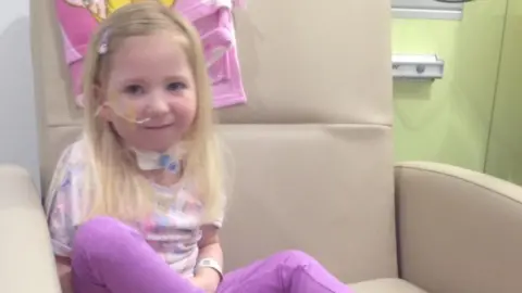Supplied Four-year-old Ava has long, light-blonde hair and is seated on a large, light‑coloured hospital-style chair. She is wearing a pastel, multi-coloured top and bright purple trousers, with legs drawn up onto the chair. A medical band is visible on Ava's wrist, and a medical device is positioned at the front of the neck area. Behind the chair, a pink blanket or towel featuring a cartoon-style illustration hangs over the backrest. The surrounding room includes light green and white walls, along with medical fixtures and equipment mounted on the wall.