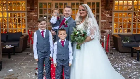 Jon/Memories Made Photography Kimberly, who is wearing a white wedding dress and veil, is posing for a photograph with her husband and three children, who are wearing matching grey suits and ties. 