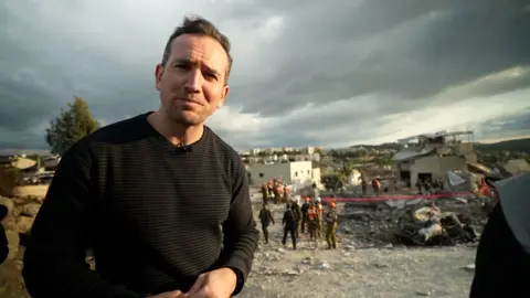 A man wears a brown jumper standing in front of rubble in Israel.