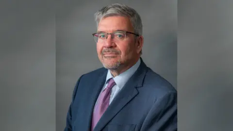 A headshot of Nick Mannion, the Labour leader of Cheshire East Council. He has short grey hair and is wearing clear glasses with a reddish brown frame, a dark blue suit, light blue shirt and purple patterned tie. 