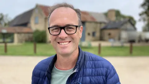 Luke Deal/BBC A head and shoulders photo Hektor Rous. He has short greying hear and black glasses. He is wearing a blue padded jacket and a light green top underneath, and he is stood in front of an old farm building