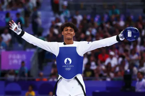 Getty Images Caden Cunningham stands pinch his arms stretched wide, a bluish helmet successful 1 hand. He is smiling and wearing each achromatic pinch a bluish vest pinch nan Paris 2024 logo on. The crowd down him is blurred.