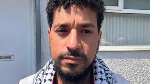 BBC/Phil Bodmer A man with dark hair and dark facial hair, with a black and white scarf. Behind him is a window into a building.