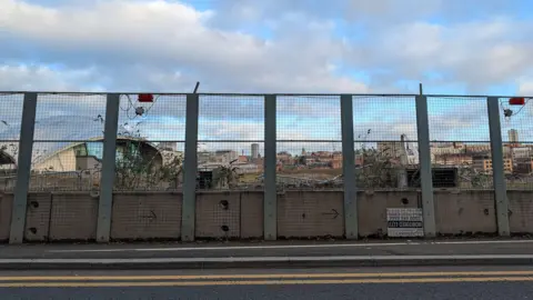 BBC A wire fence with a road in front of it. Double yellow lines are on the edge of the road. Behind the fence on the left is the glass domed roof of the Glasshouse and the top of the white curved roof of the Millennium Bridge can be seen on the right. There are overgrown plants immediately behind the fence which looks aged and run down.