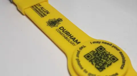 A yellow watch-style wristband which says Durham Constabulary on it, with a QR code and the words I may need your assistance.