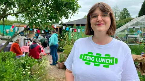A woman in a white t-shirt with a green logo saying The Big Lunch smiles. She has bobbed brown hair, fringe and glasses. Behind her are adults standing around chatting, a greenhouse, pot plants, shed and bunting in a garden space.