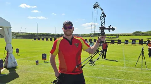 Isle of Man Archery Team Ethan Moore wearing a red T-shirt and a dark cap, as he holds up his bow. In the distance you can see a row of targets on wooden sandwich boards on a green field behind.