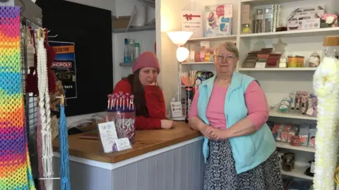 Joanna Taylor/BBC Sarah Walker standing by her shop counter, Linda Walker standing the other side of the counter surrounded by colourful gifts