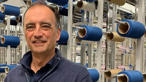 David Collinge with grey hair wearing a navy blue polo top and zip up knitted jumper standings in a textile factory with rows of large reels of blue cotton. He is smiling. 
