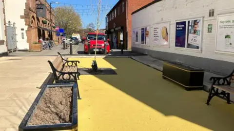 An area with benches and a large yellow section of ground. There is a car in the background and a coffee shop. It is a bright day and there is some shade from a high wall.