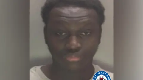 A police mugshot showing Oumar Traore in front of a neutral-coloured background. He has light facial hair and short black hair.