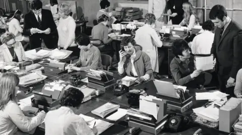 Barclays A black and white image from the 1960s showing a busy workplace of female staff answering telephones or writing on sheets of paper. Some male staff walk around overseeing the work. 