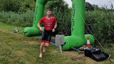 Kay Looker Matt Looker wears red and black lycra shorts and t-shirt, he runs barefoot past a large green inflatable arch on some grass.