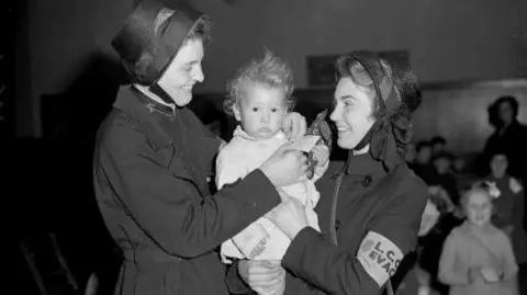 Salvation Army A black and white photo of two women in Salvation Army dress holding a toddler and smiling. Behind them are other children. 
